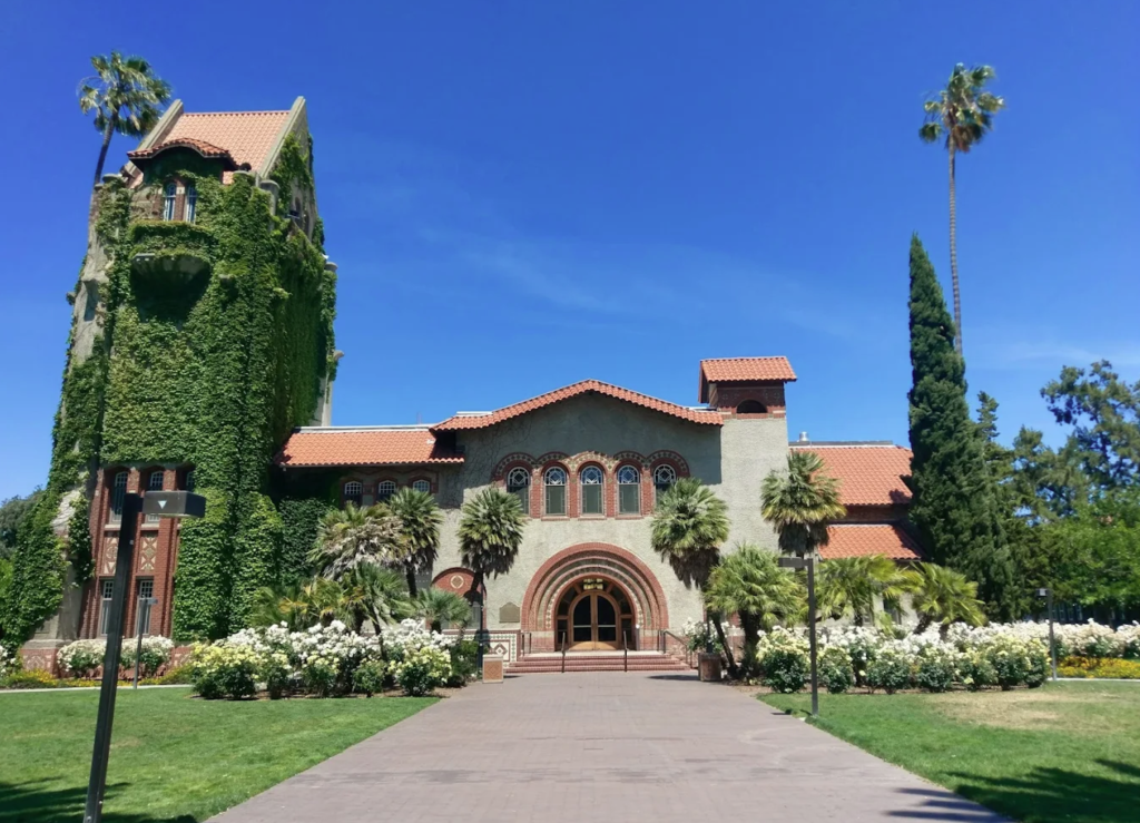 A large, old building with red-tiled roofs, a large archway entrance, and a tower covered in ivy on the left.