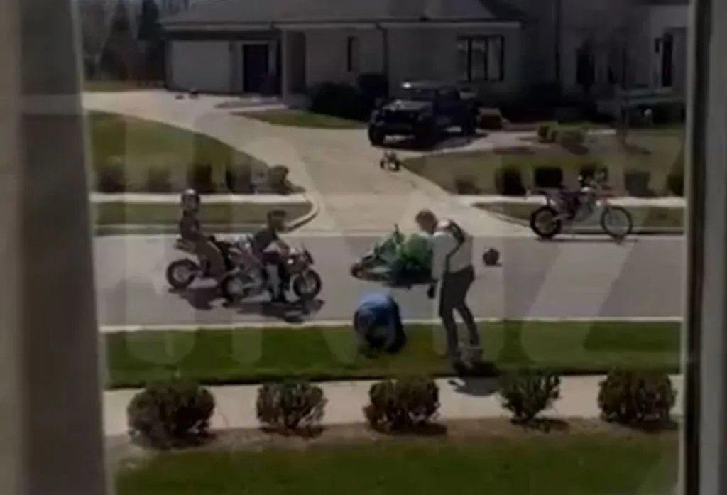 Two men, one on a small motorcycle and another standing, watch a man in blue crouch on a lawn next to a green motorcycle, with a house and driveway in the background.