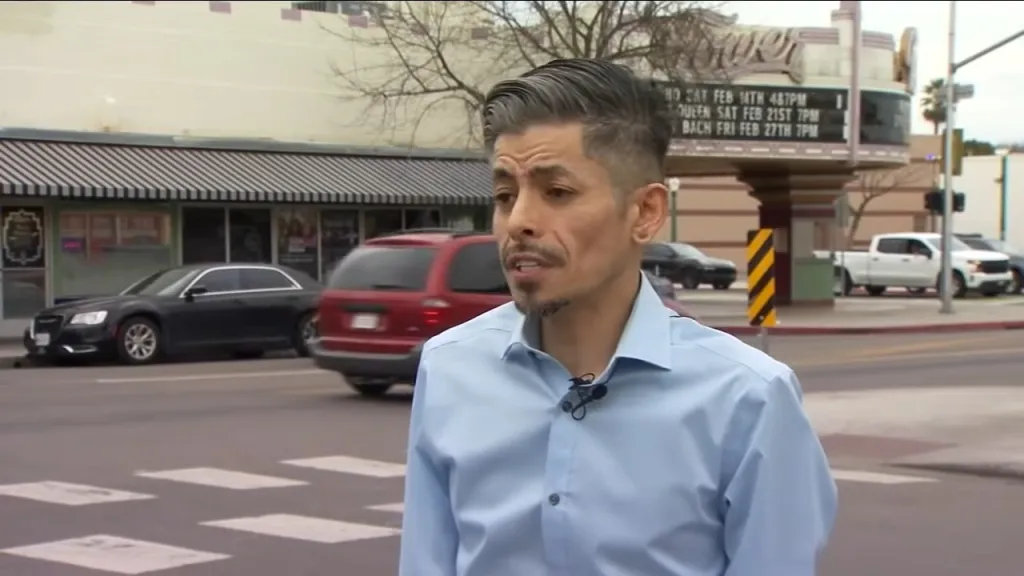 Rene Campos, a man with a goatee and slicked-back gray hair, standing in a blue button-up shirt on a city street with cars and a theater marquee in the background.