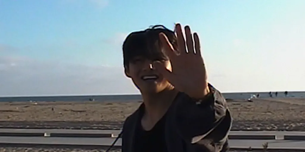 V from BTS smiling and raising his hand on a beach.