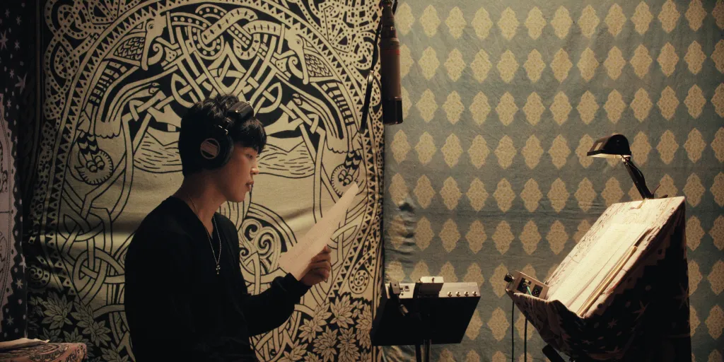 Jimin in a recording studio, wearing headphones and reading lyrics from a sheet of paper.