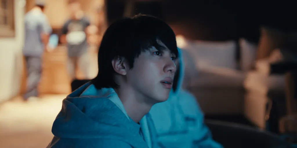 Jin from BTS looking contemplative in a blue hoodie, with other people in the blurred background.