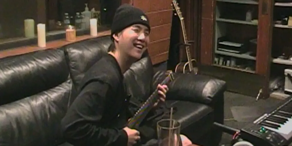 Suga of BTS laughing while playing an instrument in a recording studio.