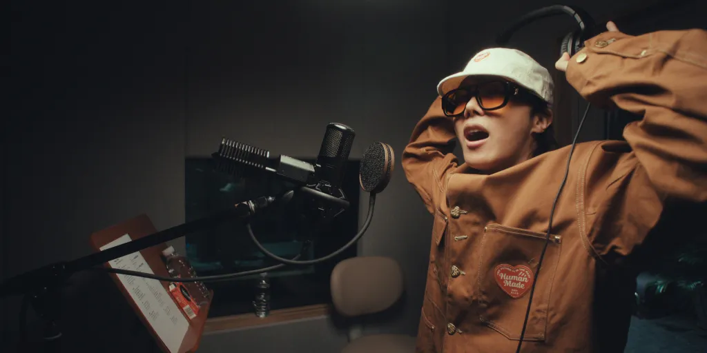 J-Hope recording vocals in a studio, wearing sunglasses and a cap, with a microphone and headphones.