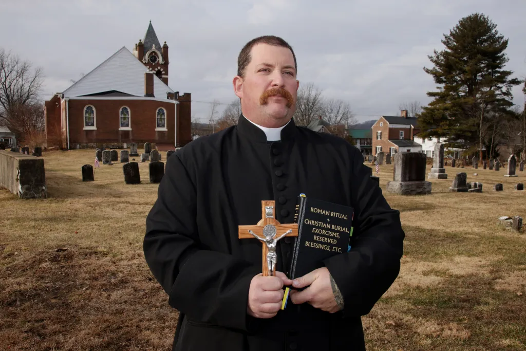 Priest dan todd standing outside a church holding a bible and crucifix looking at the camera