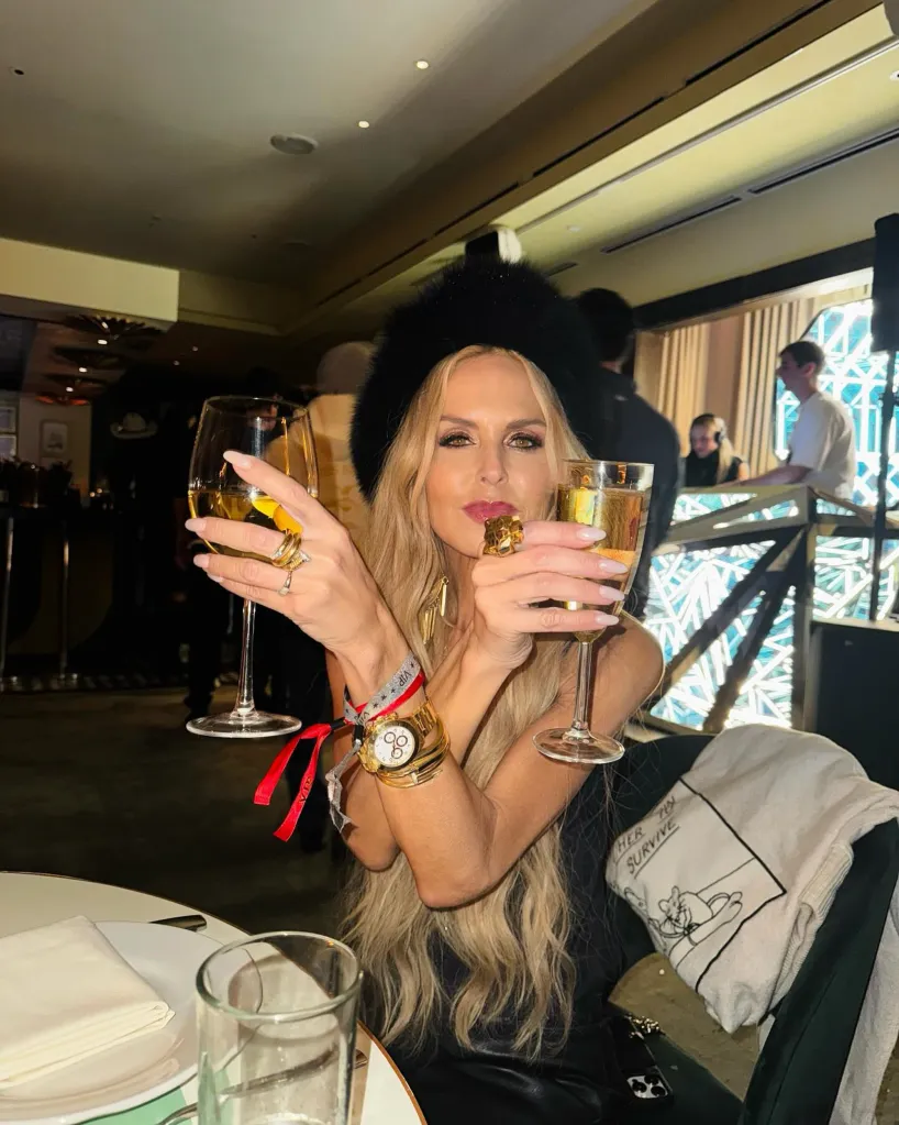 Rachel Zoe holding two glasses of champagne, wearing a black fur hat, a black outfit, and a golden watch.