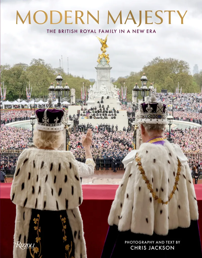 The King and Queen of England, in their crowns and fur-trimmed robes, wave to a large crowd from a balcony.