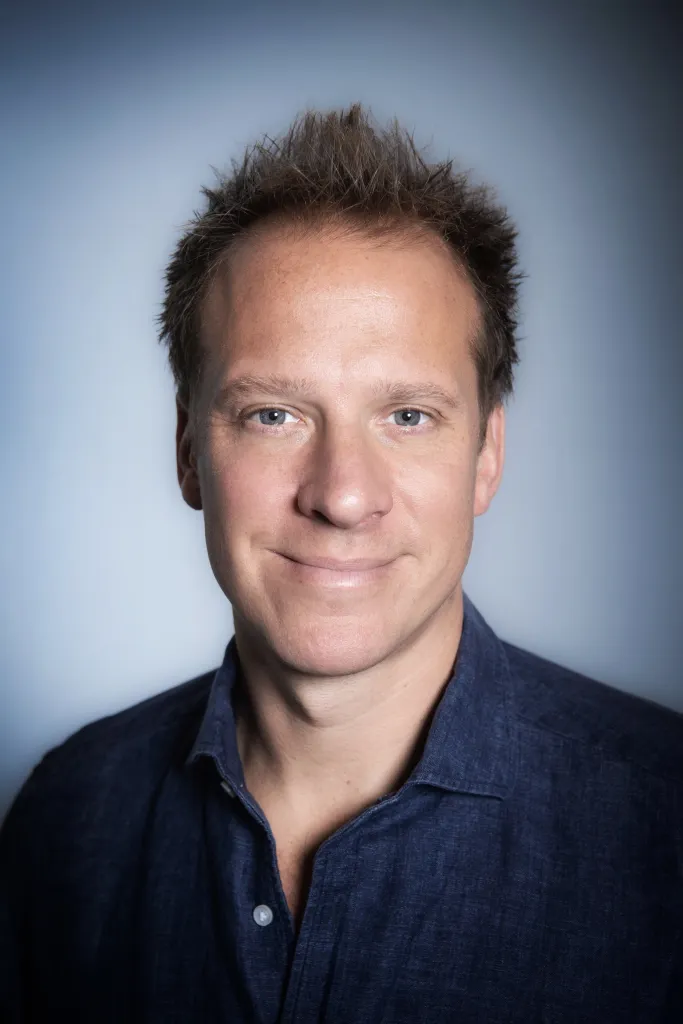 Chris Jackson, a man with light skin, short brown hair, and blue eyes, smiles faintly while wearing a dark blue collared shirt.