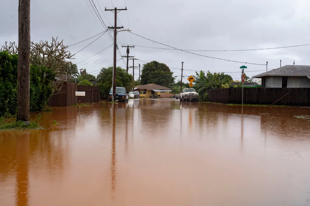 A road in Waialua, Hawaii, is covered in brown floodwaters, with cars submerged up to their tires and power lines visible overhead.