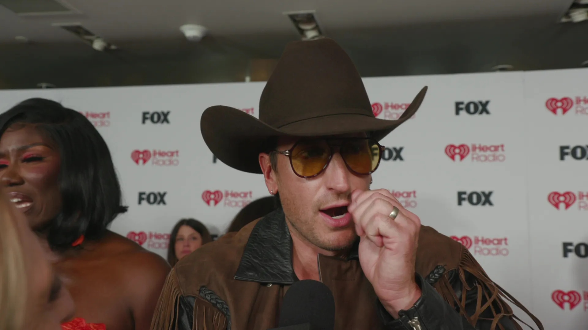 Russell Dickerson, wearing a cowboy hat and sunglasses, speaks into a microphone at an iHeart Radio and Fox event.