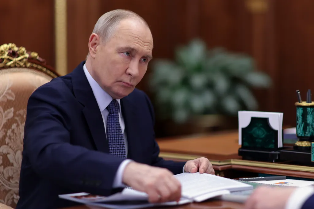 Russian President Vladimir Putin in a suit and tie, looking left while seated at a table with papers.
