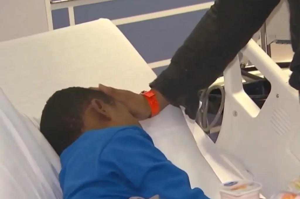 A person in a black sleeve touching the forehead of a child lying in a hospital bed.