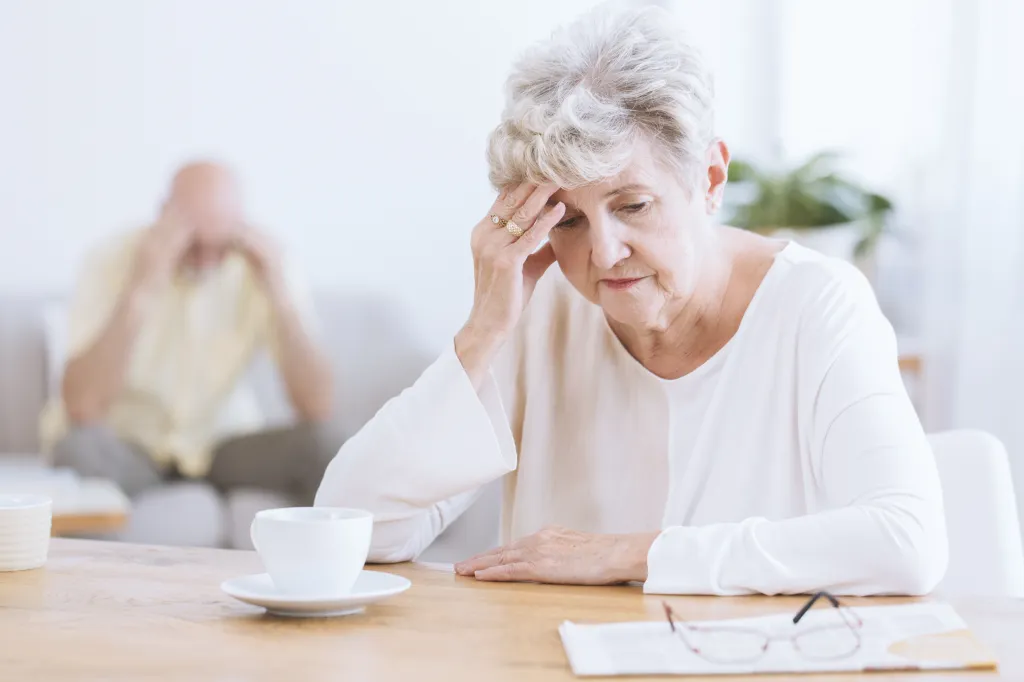 Sad senior woman sitting at a table with her hand on her forehead after a quarrel, with her husband in the background.