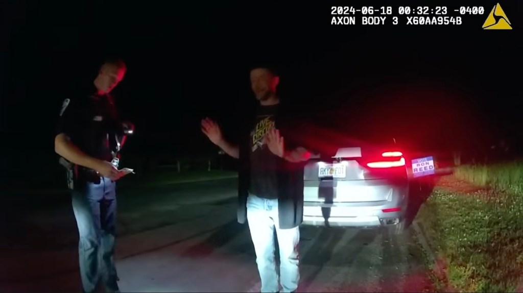 Justin Timberlake standing with his hands raised during a DWI stop, with a police officer and the back of a car visible at night.