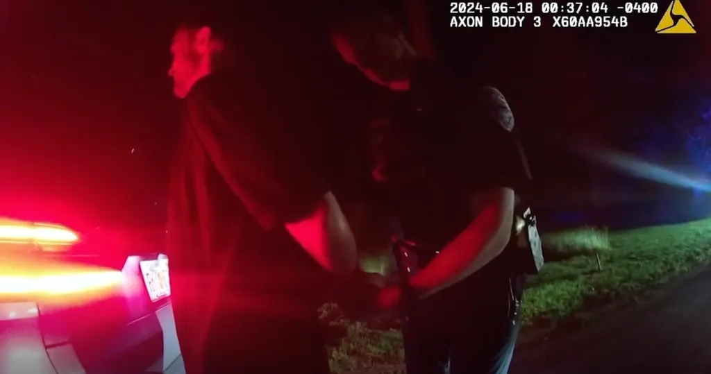 Bodycam footage of a police officer handcuffing a person at night with the flashing red light of a police car visible.