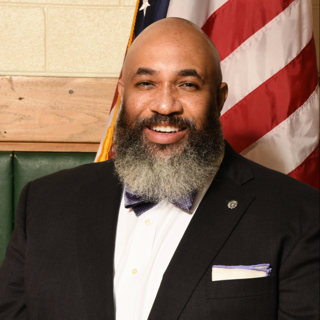 Photo of Dr. Corey W. Reynolds, a man with a bald head, dark brown skin, and a large gray beard, smiling in front of an American flag.