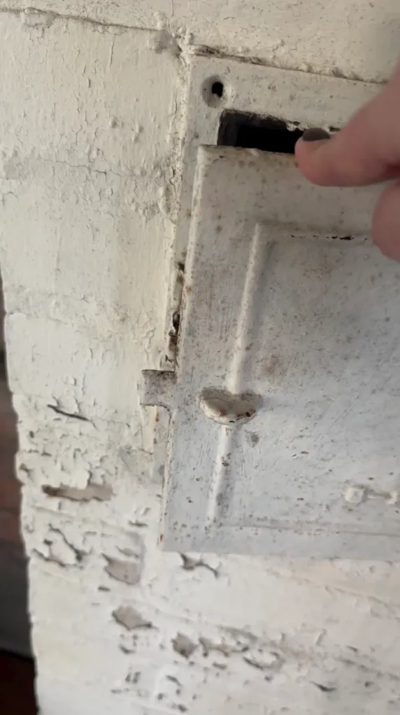 A hand opening an old, white, rusted metal door on a wall with peeling paint.