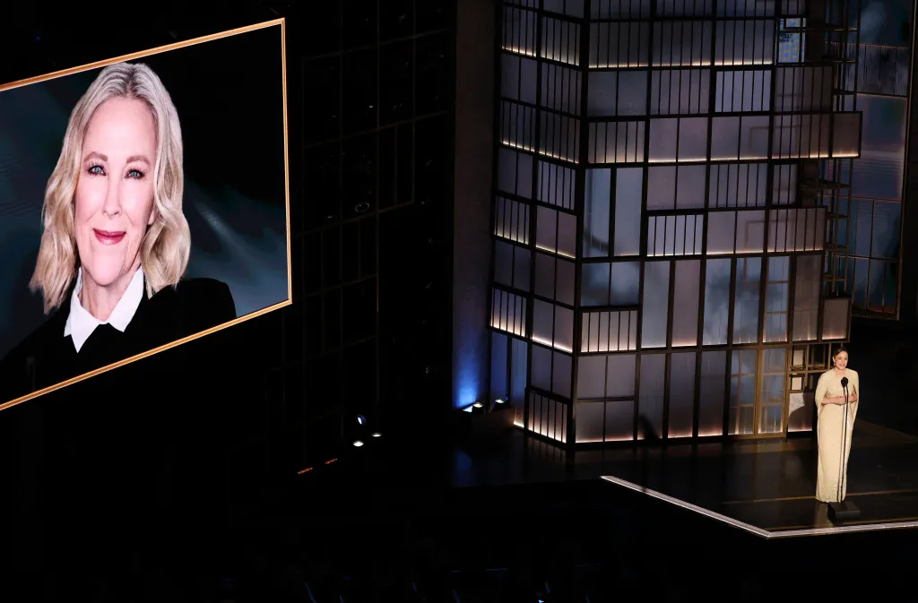 Rachel McAdams speaks on stage during an in memoriam segment at the Academy Awards, with a screen displaying a photo of Catherine O'Hara.