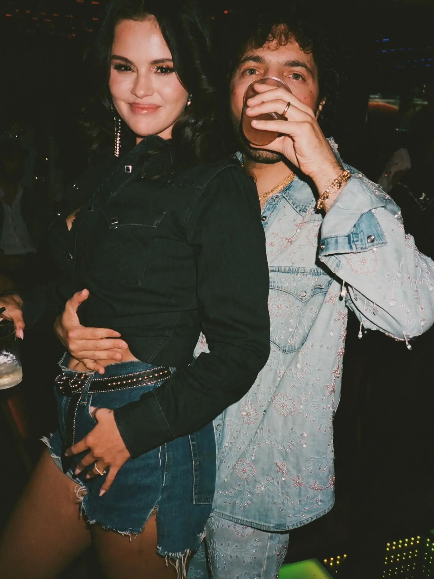 Selena Gomez in denim shorts and a dark denim top, embracing Benny Blanco who is drinking from a glass, both wearing denim outfits.