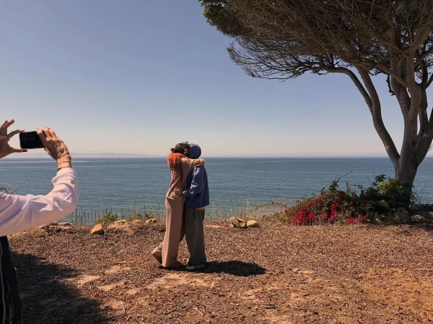 Selena Gomez and Benny Blanco embracing by the ocean, with someone taking a photo of them.