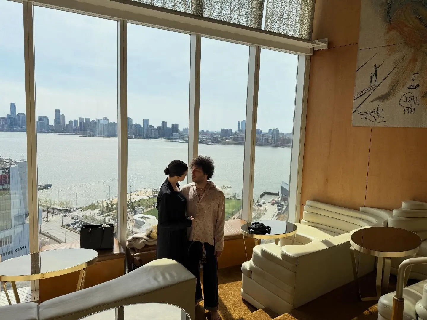 Selena Gomez and Benny Blanco gazing at each other in a sun-filled room with a city and river view.