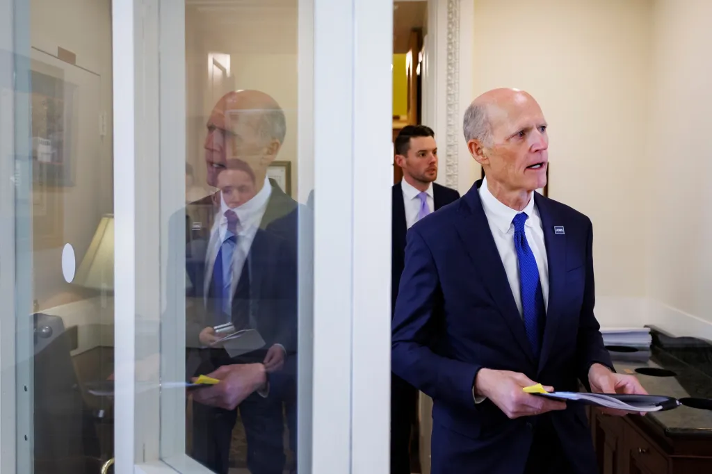 Sen. Rick Scott walking with documents in hand, with another man in a suit walking behind him.
