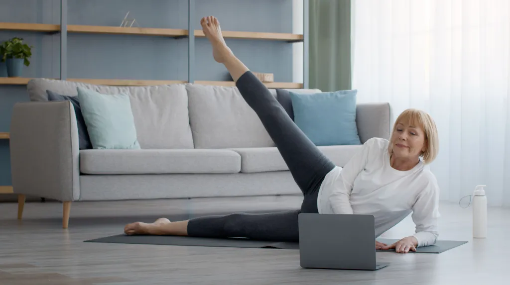 A senior woman doing leg raises on a yoga mat in front of a laptop.