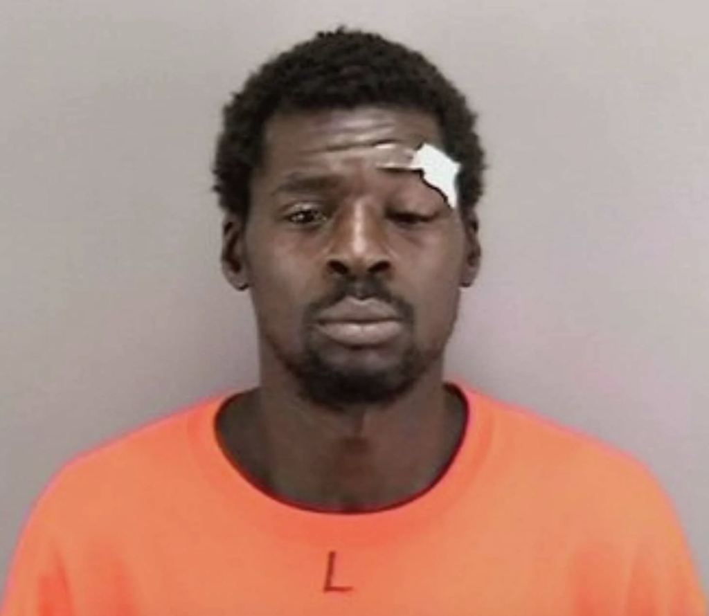 Mugshot of a Black man with a bandaged forehead and swollen left eye, wearing an orange shirt.
