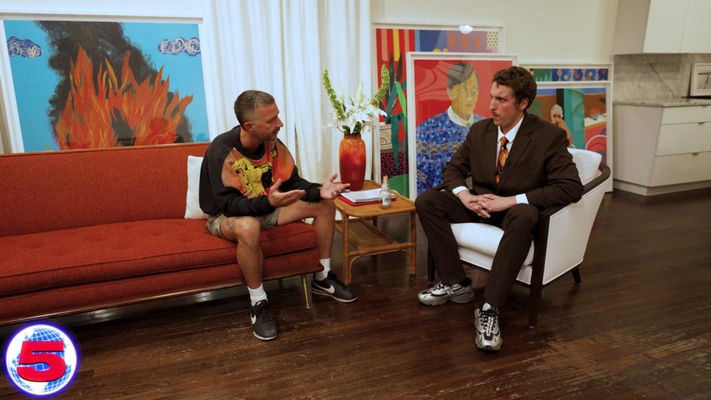 Shia LaBeouf, in a black sweatshirt and shorts, being interviewed by Andrew Callaghan, in a black suit and orange tie and sneakers.