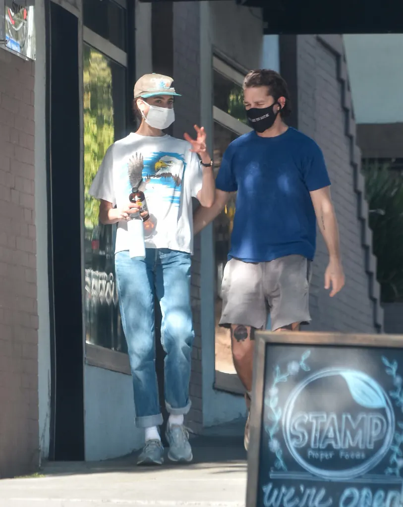 Shia LaBeouf, in a blue t-shirt and khaki pants, and Margaret Qualley, in a white t-shirt and blue jeans and white baseball cap, walking together, both wearing face masks.