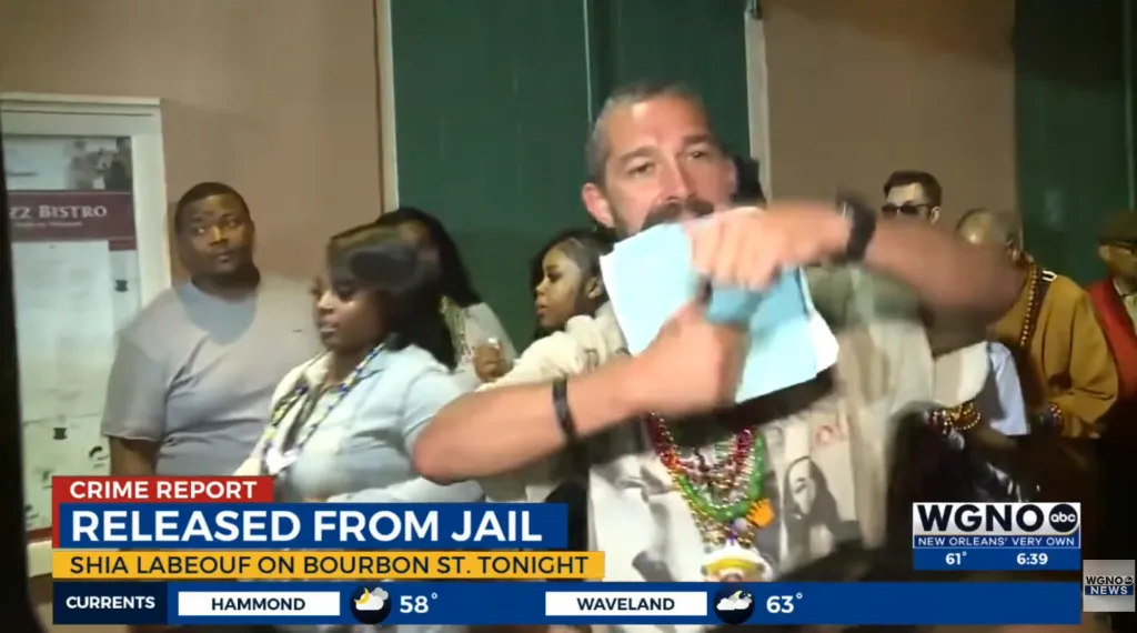 Shia LaBeouf, in a cream t-shirt, dancing and holding jail paperwork in his mouth.