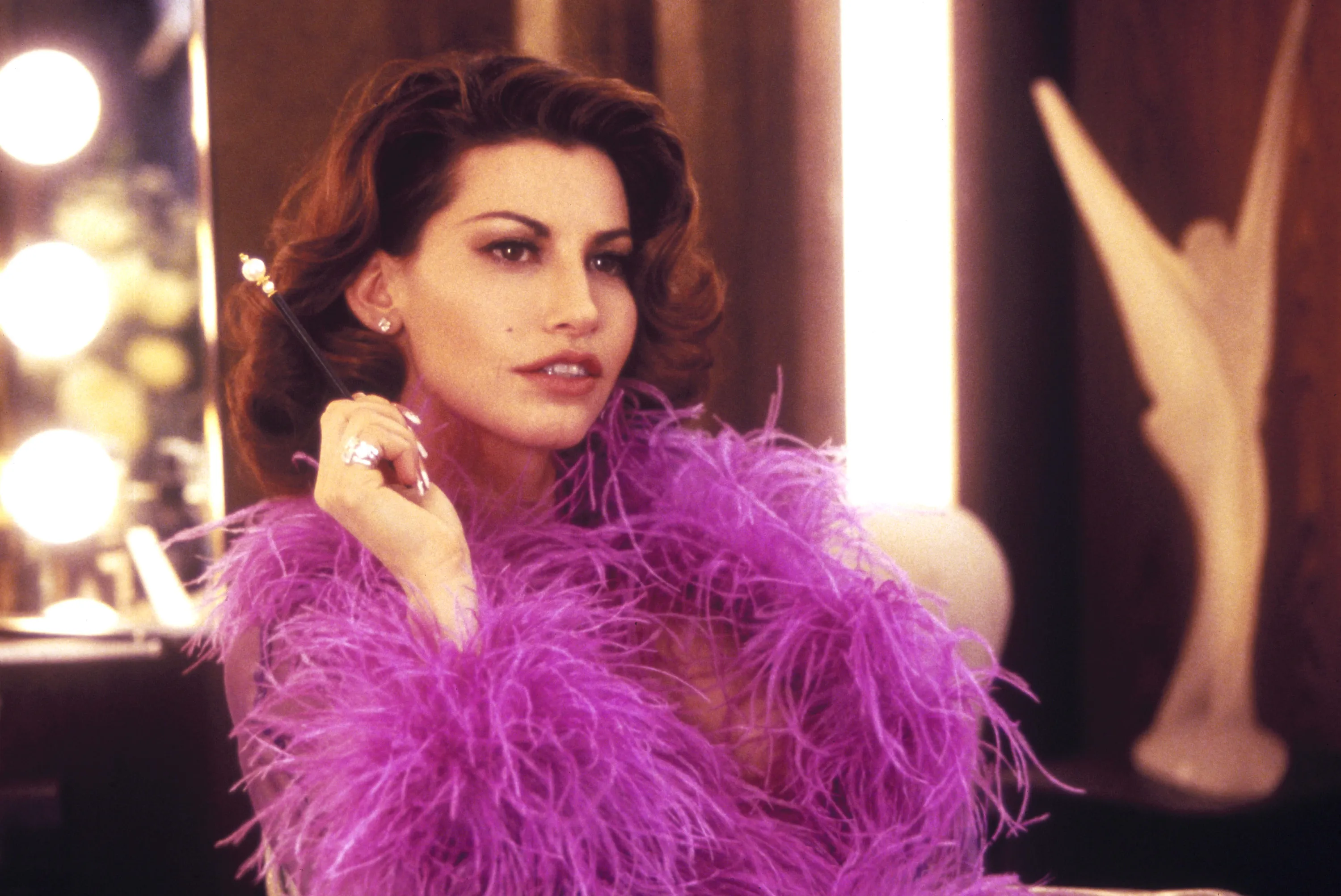Gina Gershon as a showgirl in a pink feather boa, holding a long cigarette holder.