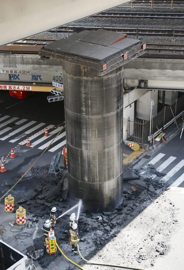 A giant pipe emerging from the ground at a sewer construction site in Osaka, with workers spraying water onto it.
