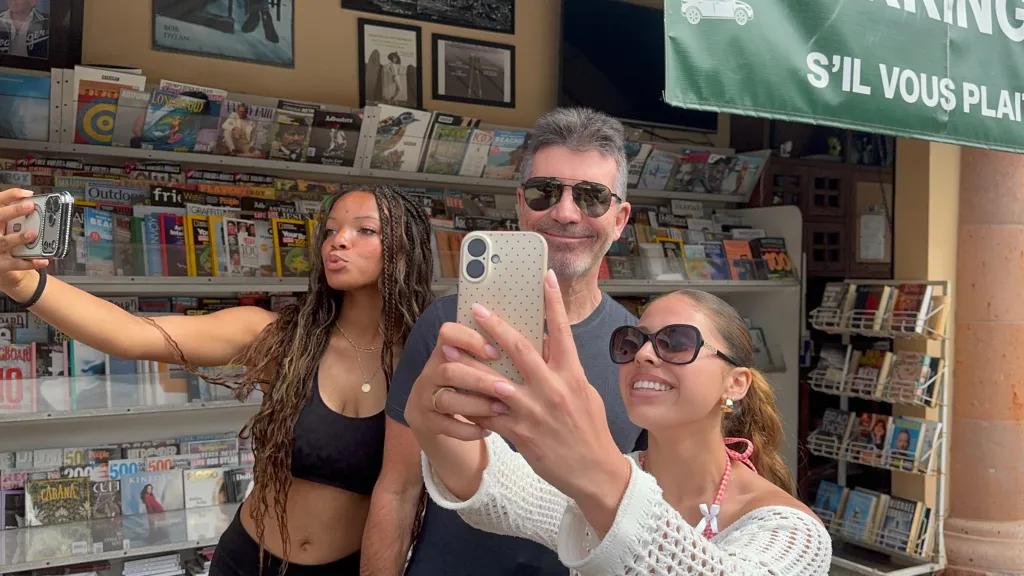 Simon Cowell takes a selfie with two women in front of a newsstand.