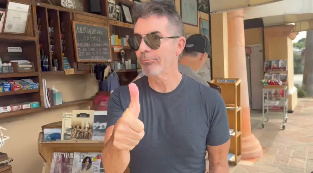 Simon Cowell posing with a thumbs up in front of a newsstand.