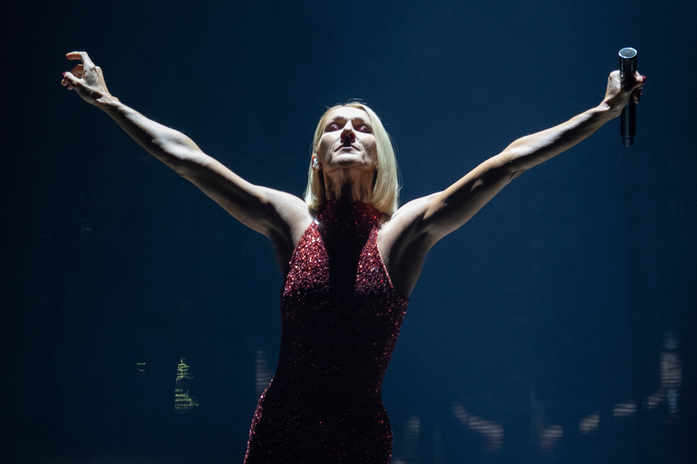 Celine Dion performing on stage in a sparkly red dress with her arms outstretched, one holding a microphone.