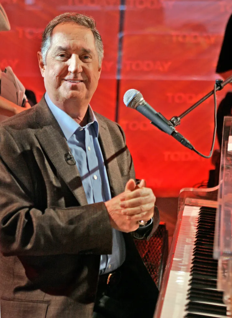 Neil Sedaka smiling with his hands clasped, seated at a piano with a microphone in front of him.