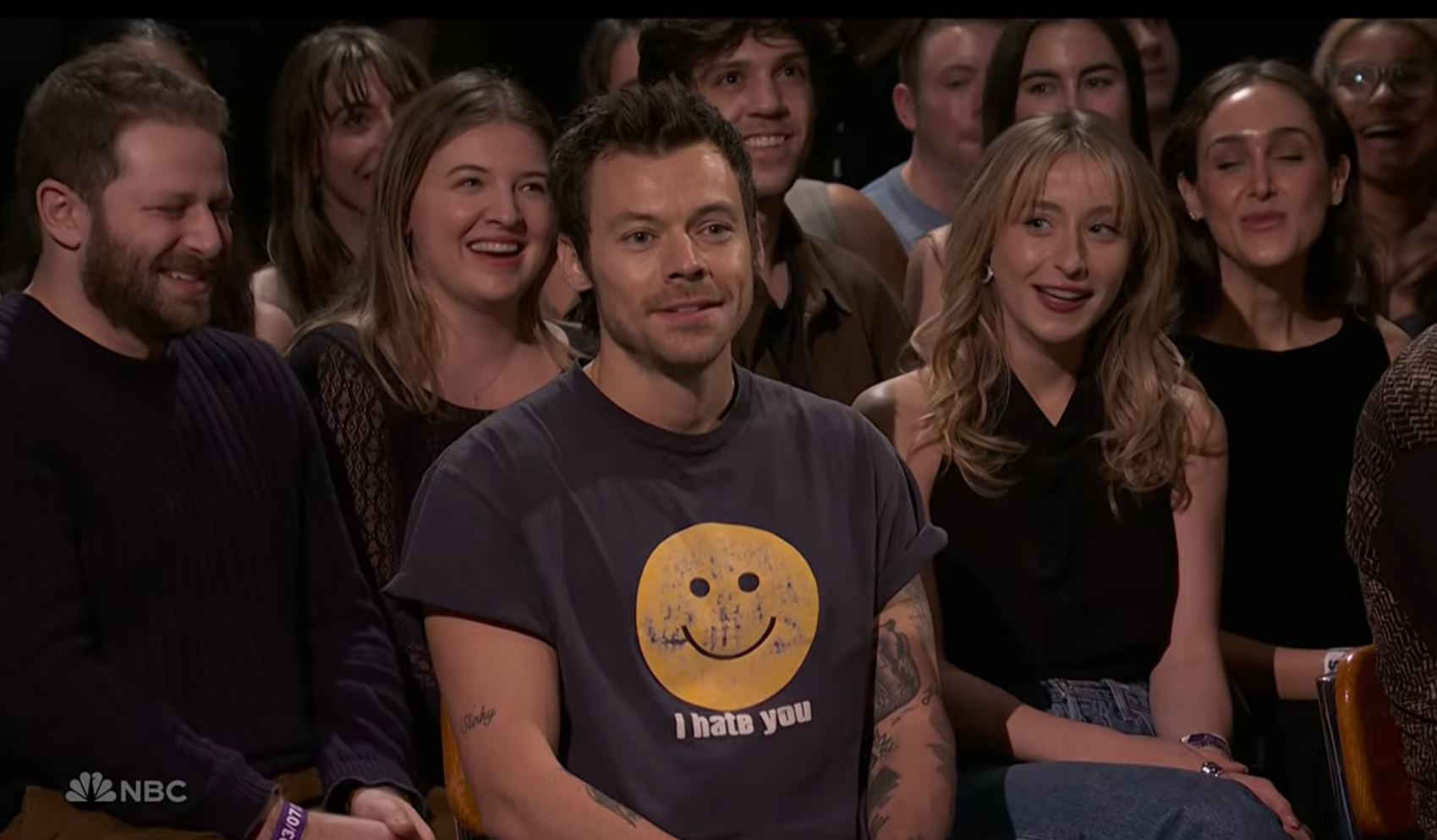 Harry Styles smiling while sitting in an audience.