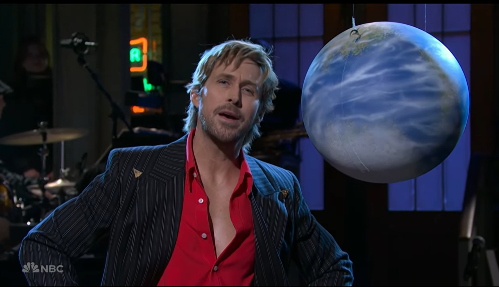 Ryan Gosling on SNL, wearing a red shirt and striped jacket, with a globe prop beside him.