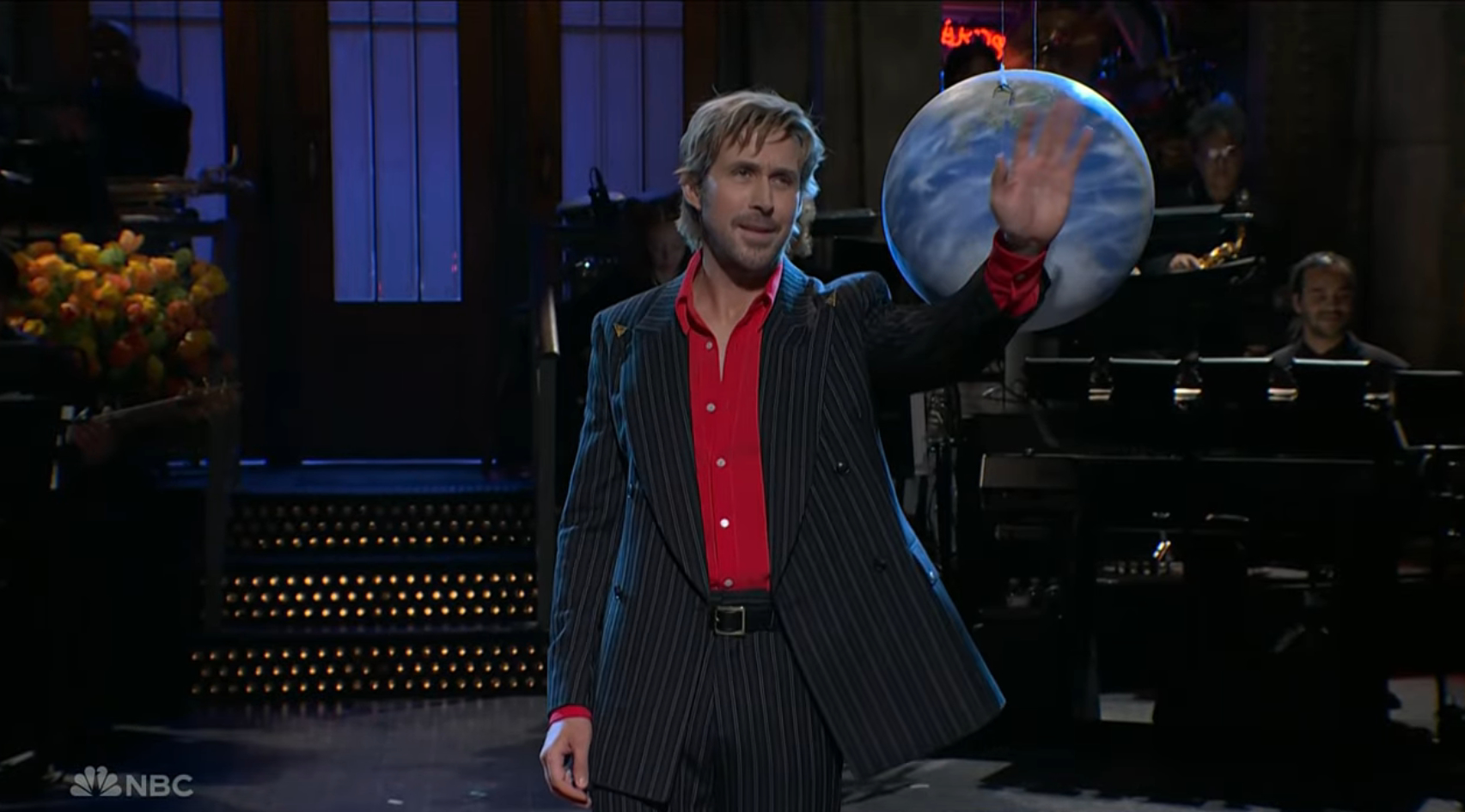 Ryan Gosling on SNL stage, wearing a pinstriped suit and red shirt, waving at a globe prop.