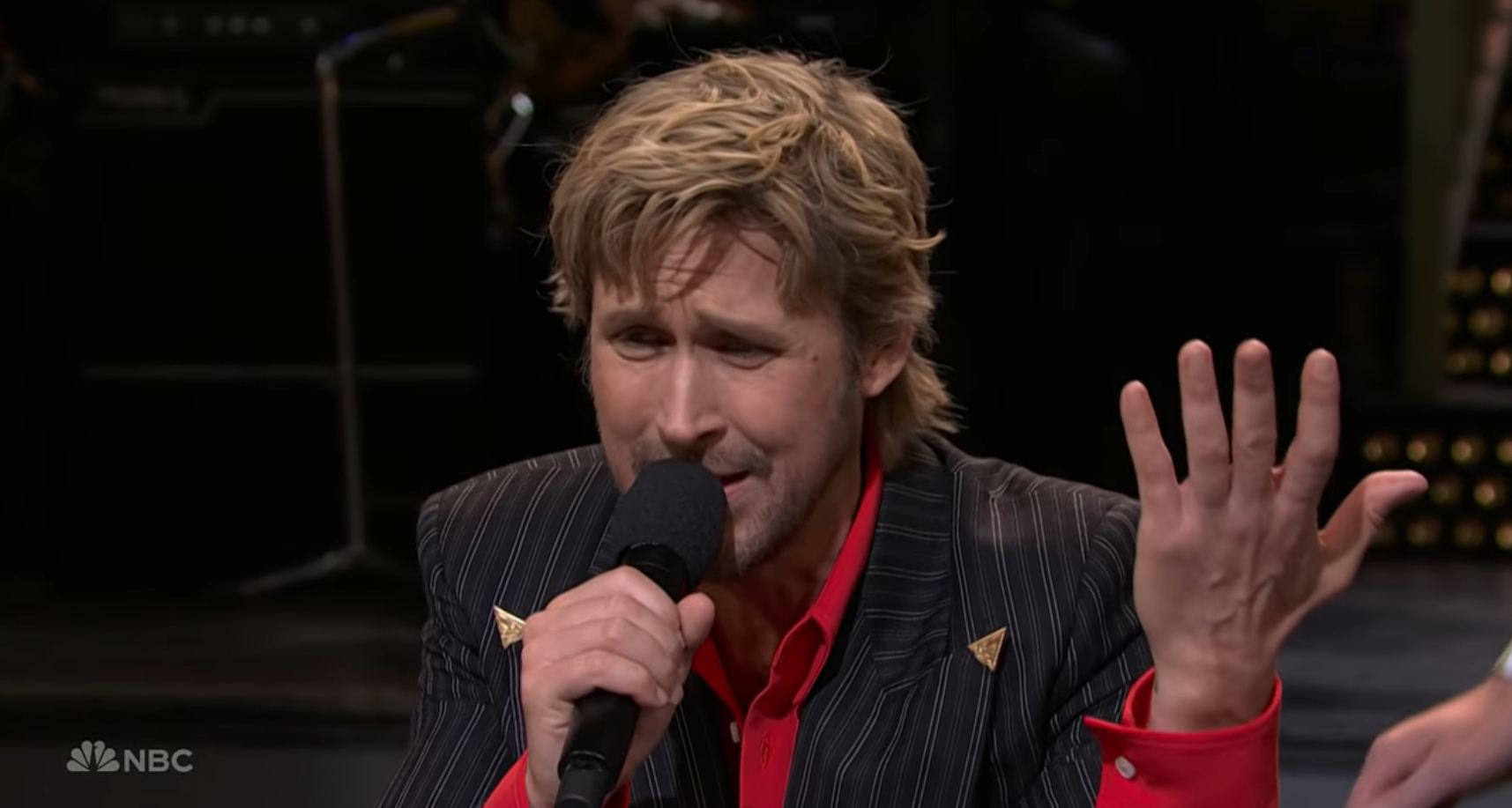 Ryan Gosling holding a microphone and raising his left hand, wearing a striped suit jacket and a red shirt.