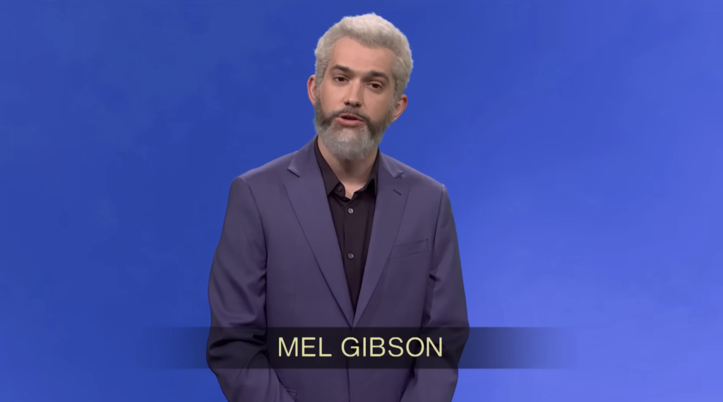 A man portraying Mel Gibson, with a grey beard and hair, stands in a blue suit against a blue background.