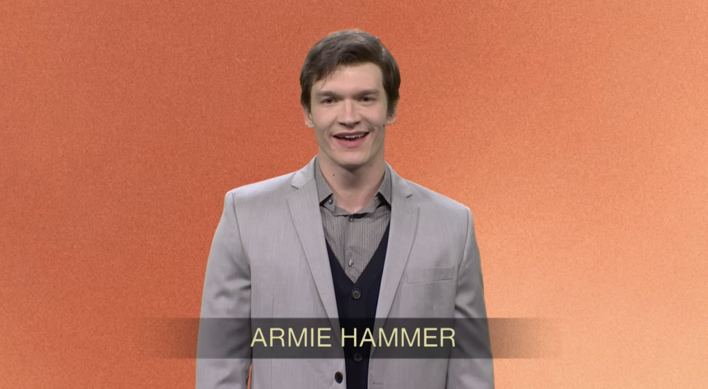 A man in a light grey suit jacket and dark vest smiling at the camera, with a title that reads 