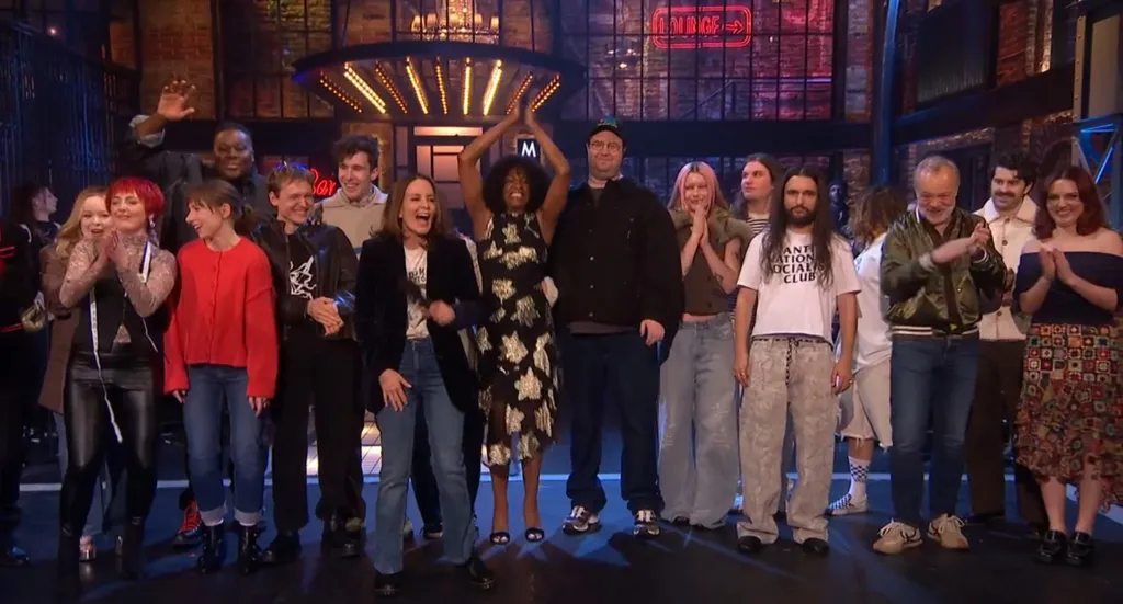 The cast of Saturday Night Live UK stands together on stage, some waving and clapping.
