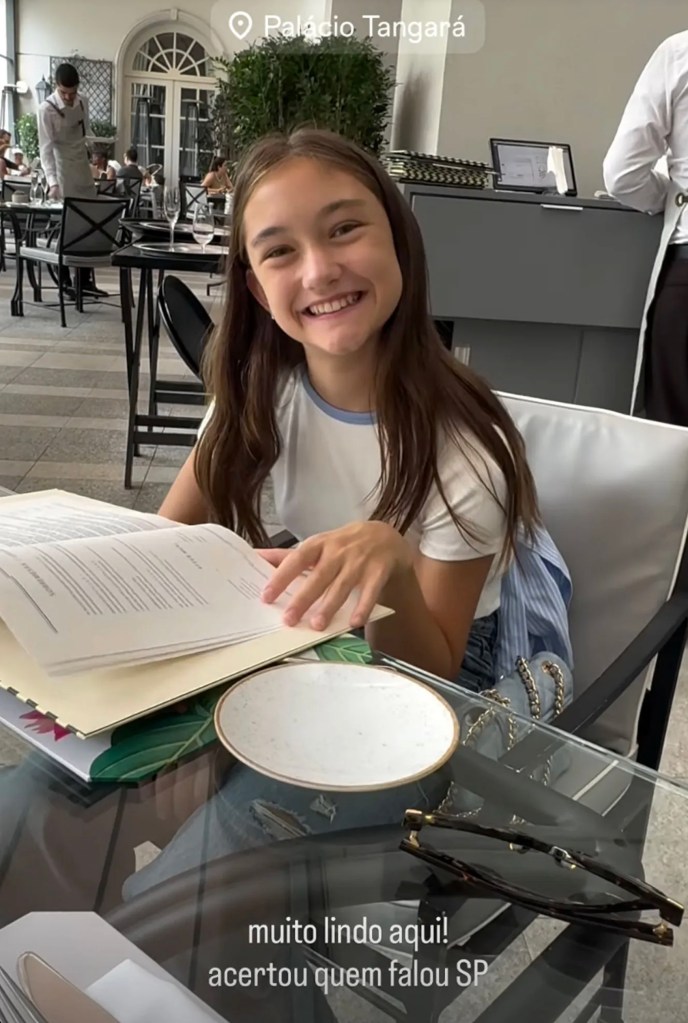 A young girl smiling and looking at the camera while holding a menu at the Palácio Tangará restaurant.
