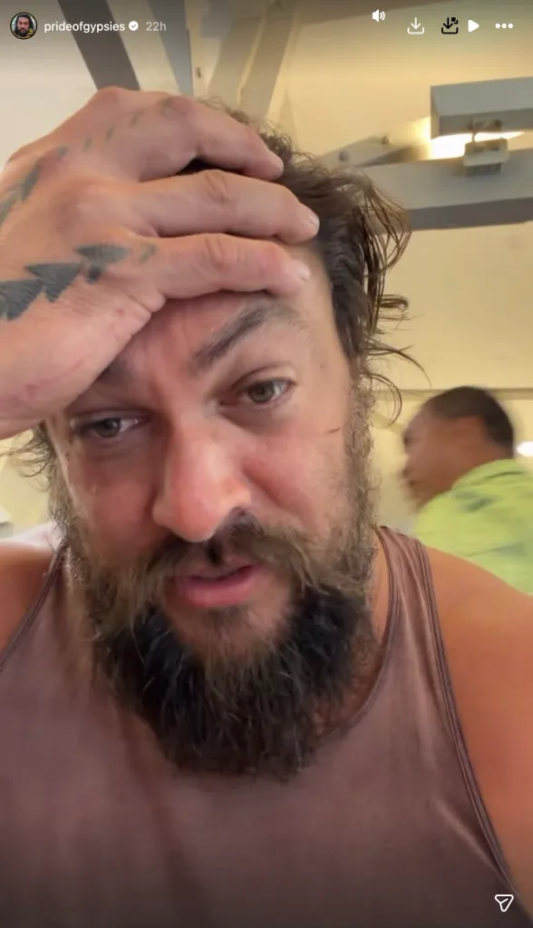 Jason Momoa in a brown tank top, with a solemn expression, holding his hand to his forehead.