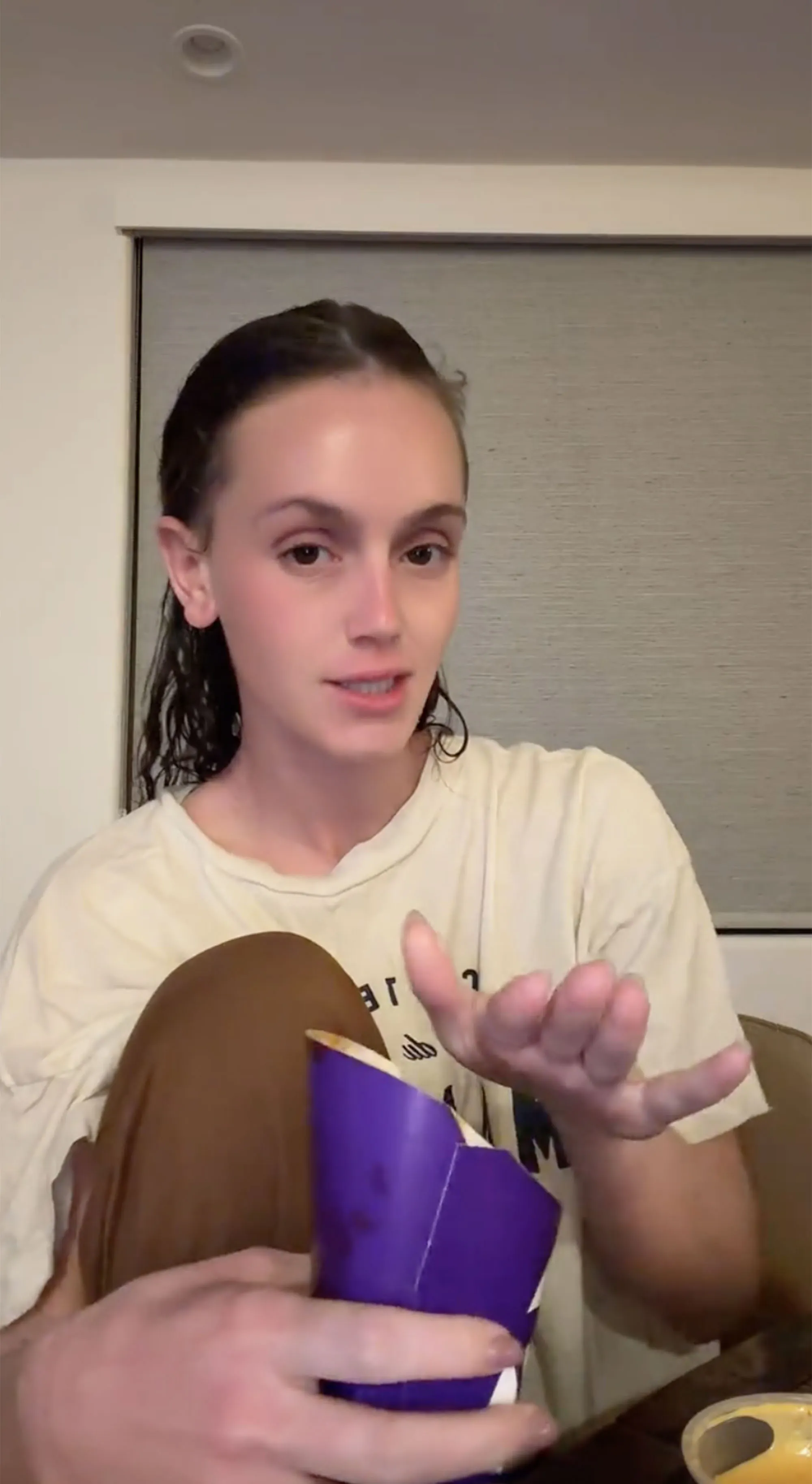 A woman sits cross-legged, holding an open purple Taco Bell container, gesturing with her other hand.