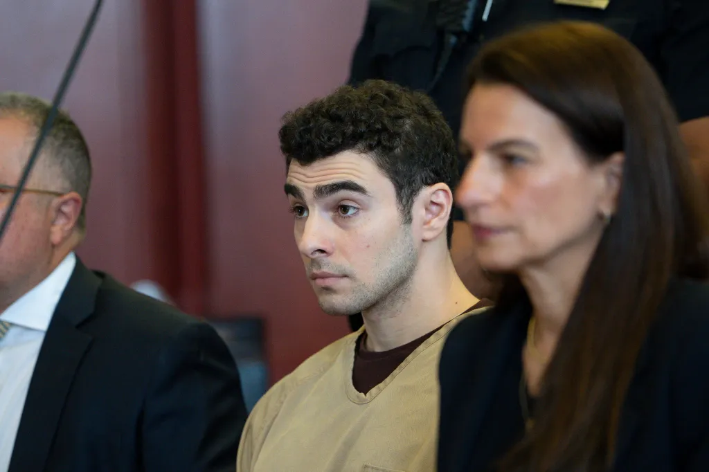 Luigi Mangione, accused of murder, sits in court wearing a tan and brown shirt.