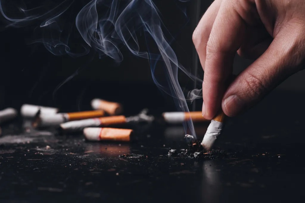 Hand crushing a lit cigarette with smoke rising, surrounded by extinguished cigarettes.