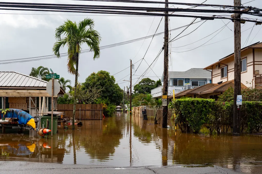 Floodwaters cover a street in Haleiwa, Hawaii, obscuring a portion of a house, power poles, and palm trees.